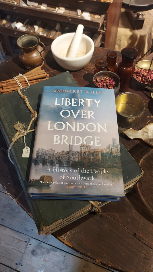 A copy of Liberty over London Bridge placed on a stack of old books wrapped in string, next to apothecary items, such as a mortar and pestle, jars, scales and dried petals