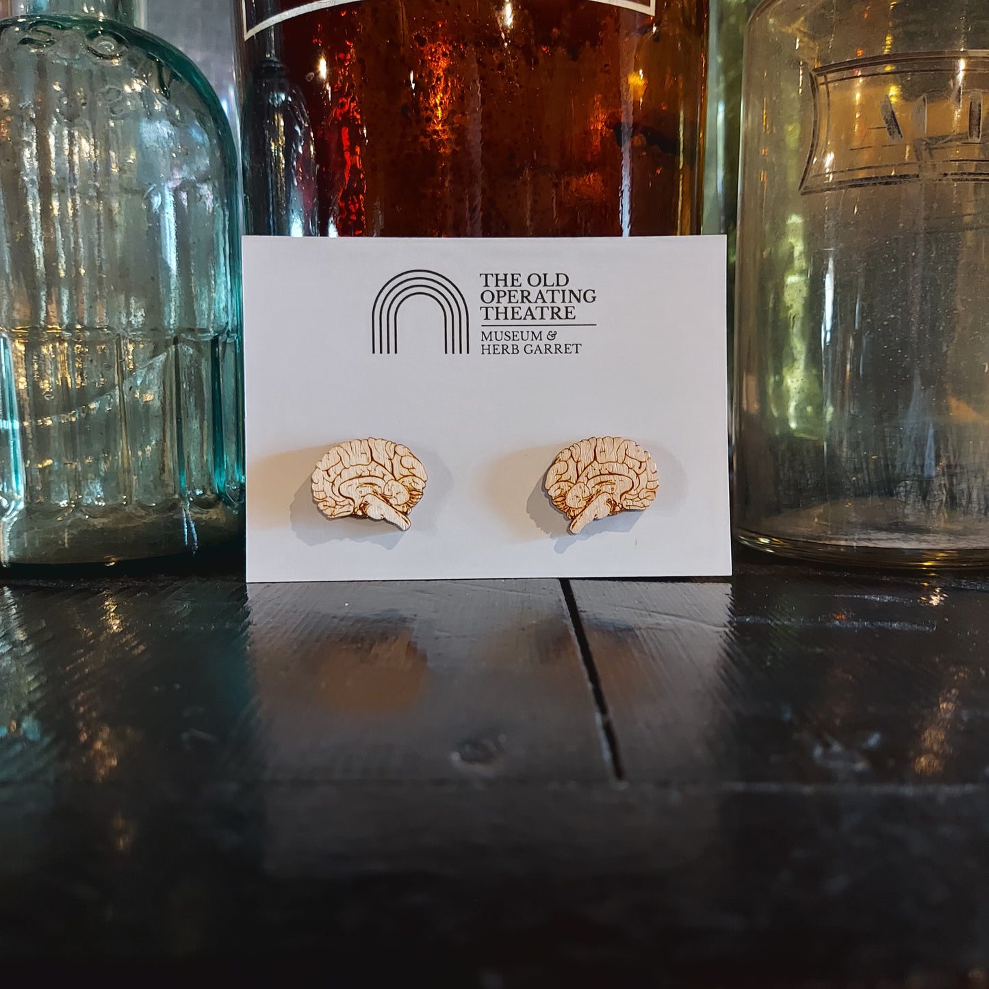 Two wooden earrings in the shape of a brain, attached to white card with the museum's logo, placed again old glass medicine bottles