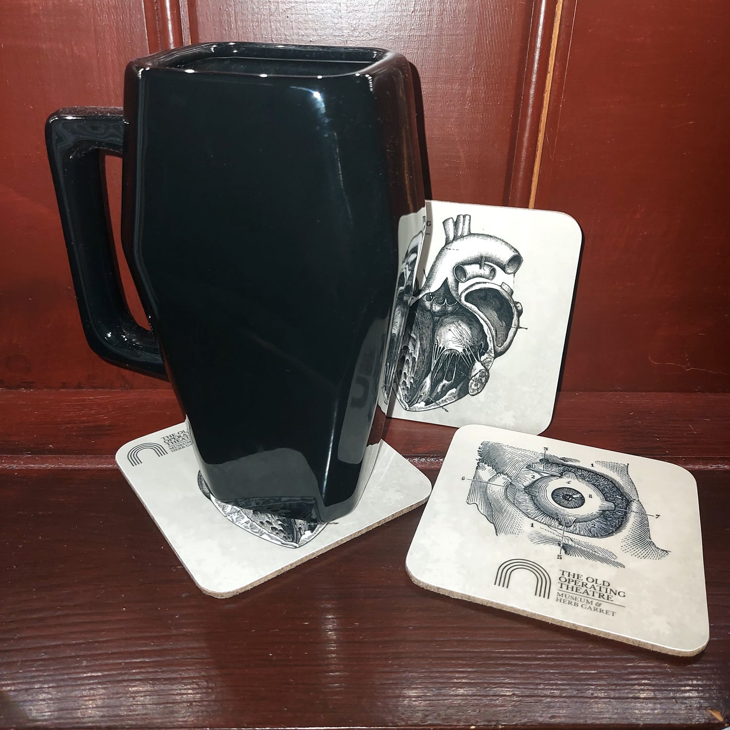 A tall black mug on a white coaster, next to two other coasters, one featuring an anatomical drawing of an eye and one a drawing of a heart