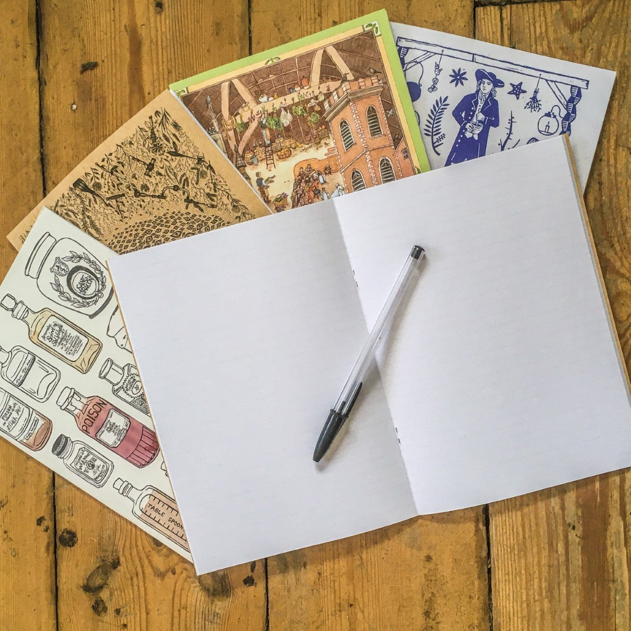 4 notebooks on wooden background, with 1 open notebook and black pen on top.