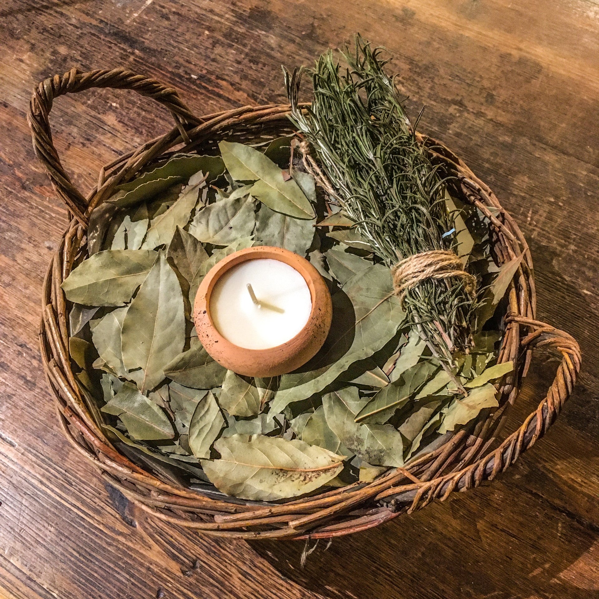 A terracotta candle placed in a basket, on top of a pile of bay leaves and a bundle of rosemary sprigs tied with brown string