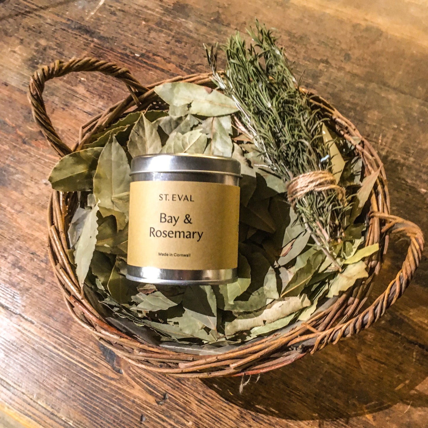 A bay and rosemary candle tin placed in a basket, on top of a pile of bay leaves and a bundle of rosemary sprigs tied with brown string