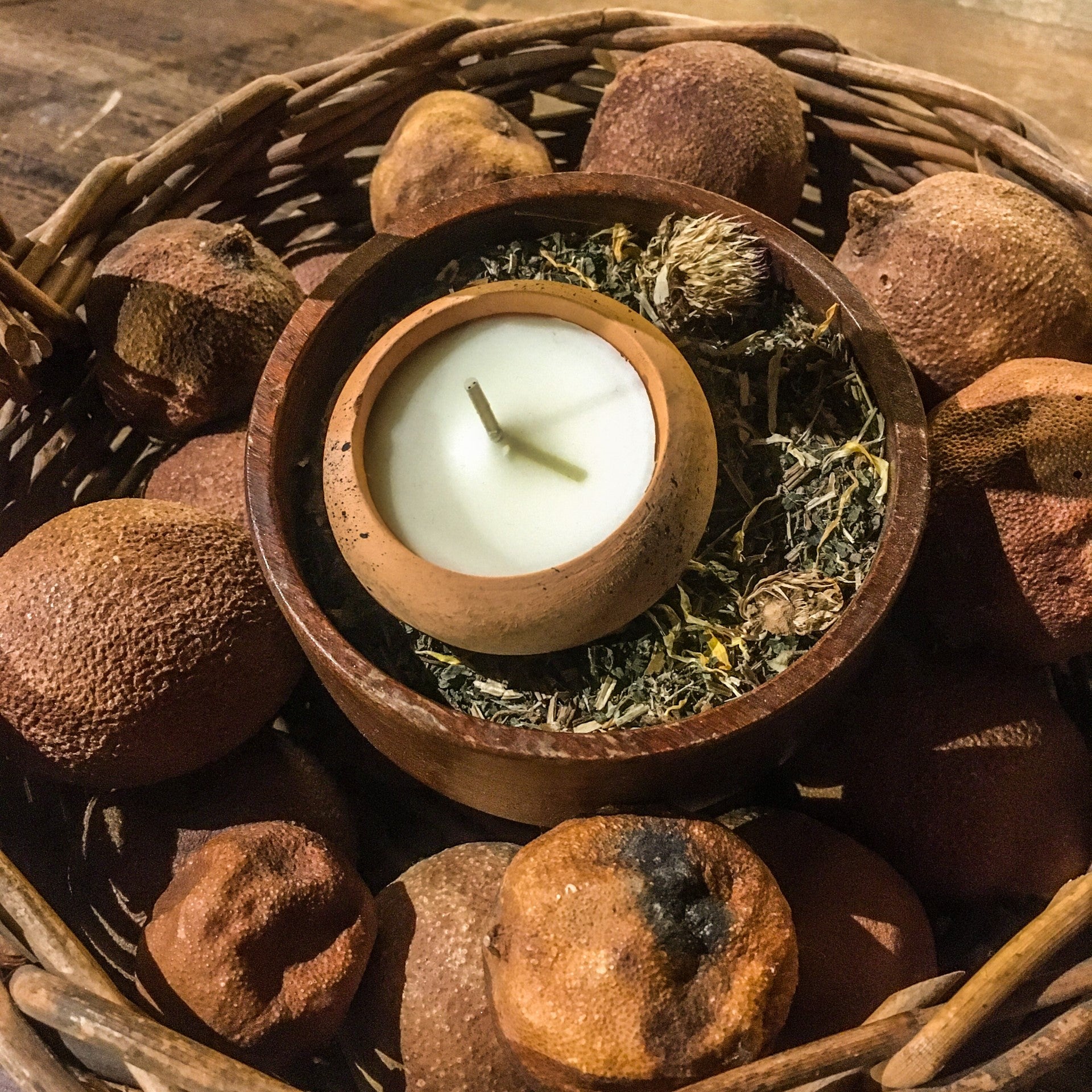 A terracotta candle placed within a wooden bowl of herbs, which sits on a wicker basket of dried fruit