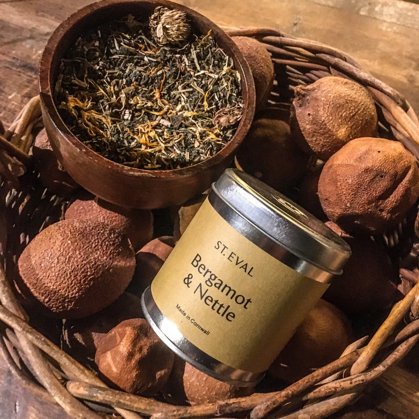 A bergamot and nettle candle tin in a wicker basket of dried fruit next to a wooden bowl of dried herbs