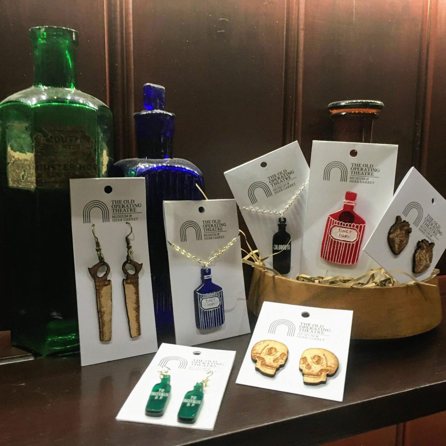 A selection of jewellery, including bonesaw earrings, apothecary bottle necklaces, skull and heart earrings, and bottle earrings, placed in front of blue, green and amber glass bottles