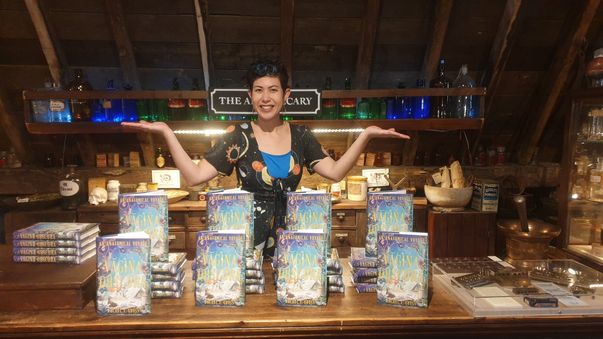 A woman stands behind the apothecary counter with her arms out to her sides; on the counter are multiple copies of the book Vagina Obscura