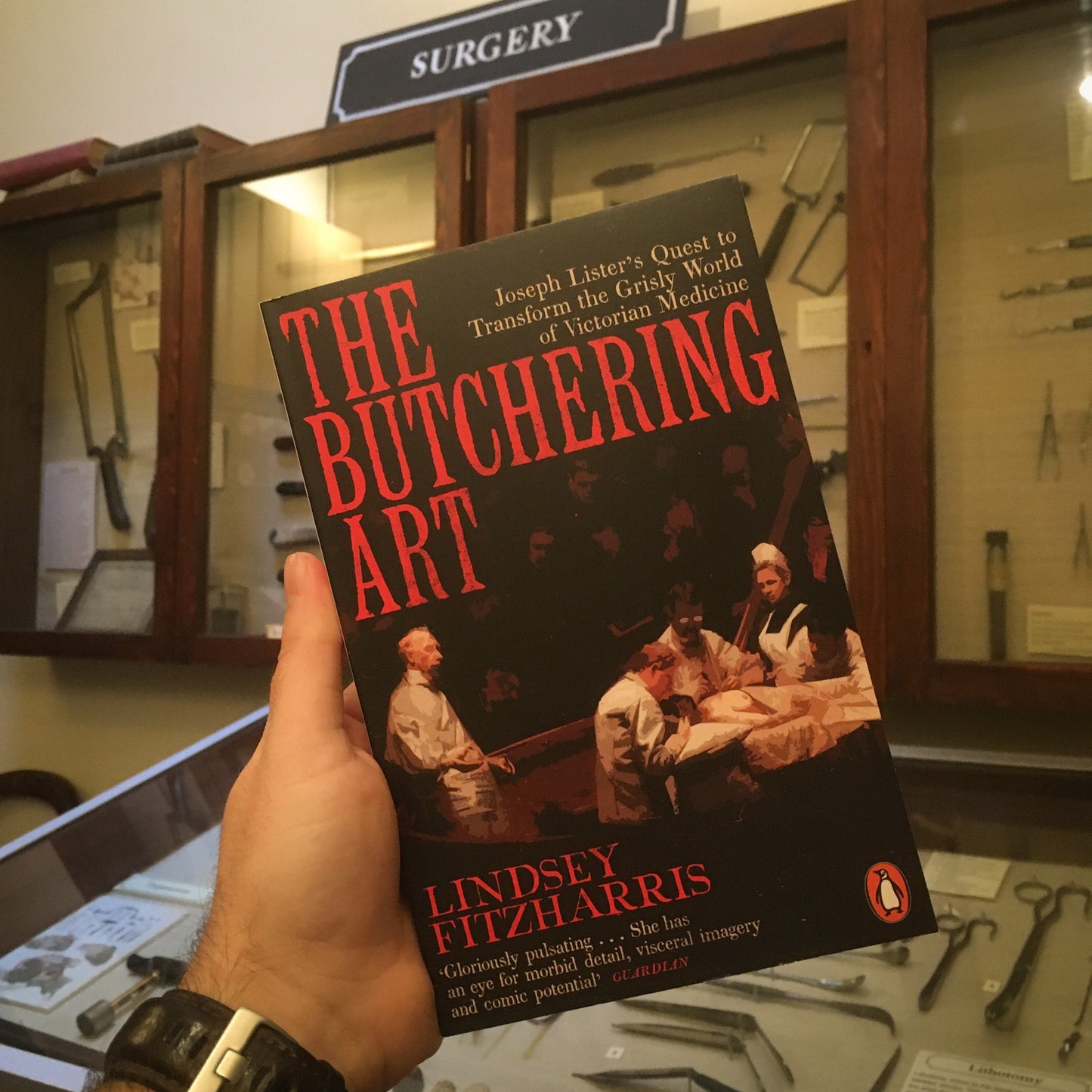 The book The Butchering Art held up in from of the museum's collection of surgical instruments