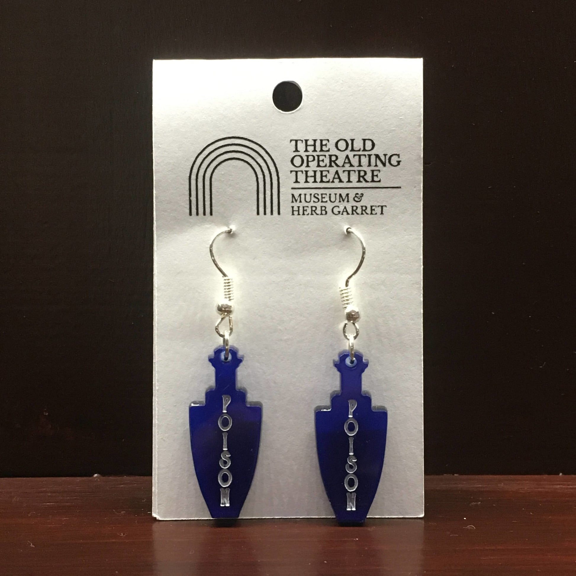 Pair of blue dangle earrings in the shape of a bottle. 'Poison' is written vertically on the bottle.