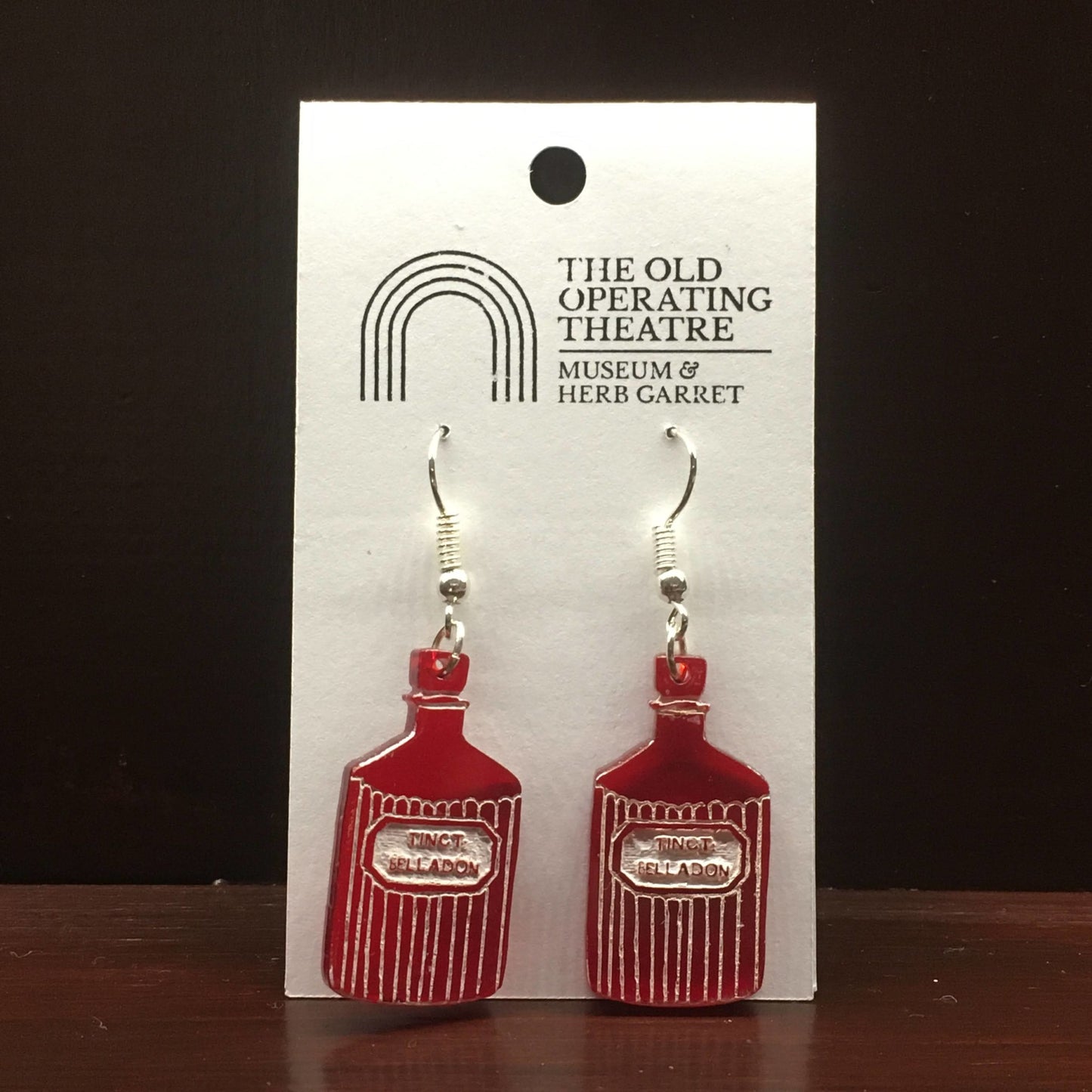 Pair of red dangle earrings in the shape of a bottle with the label 'tinct. belladon'