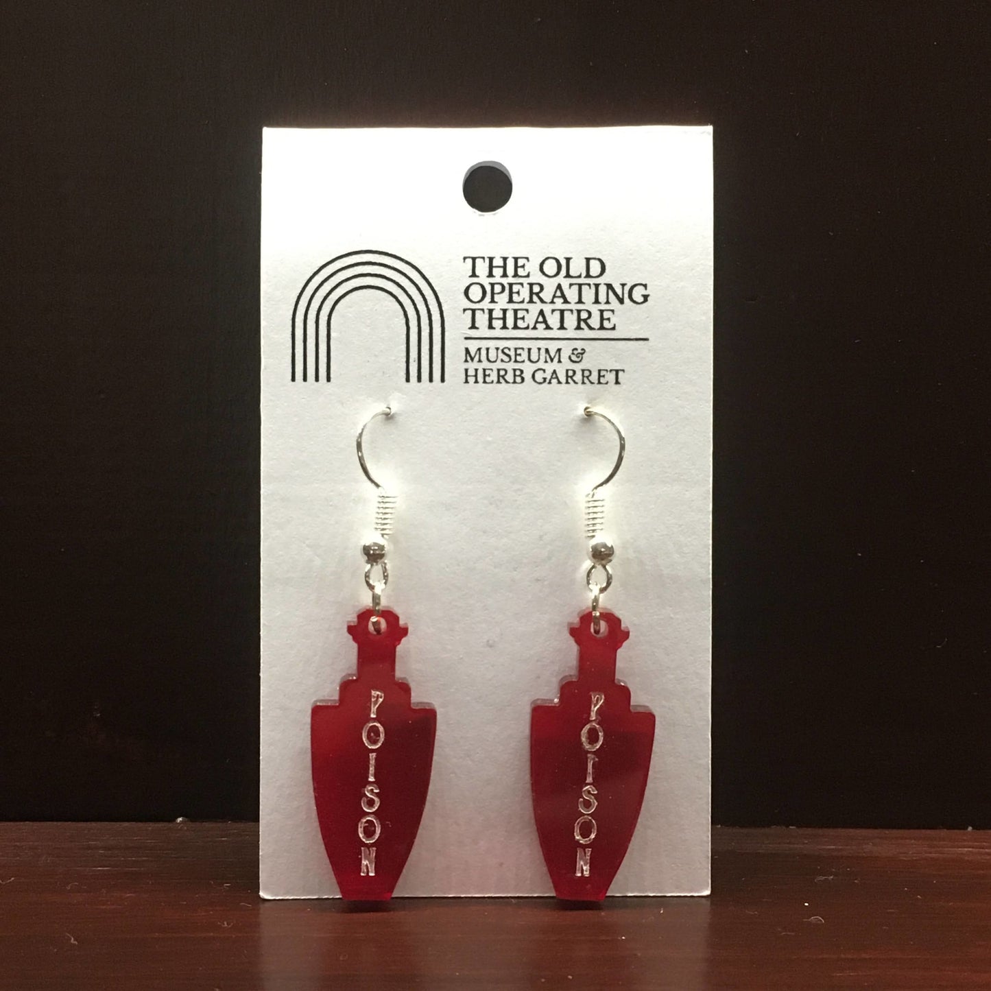 Pair of red dangle earrings in the shape of a bottle. 'Poison' is written vertically on the bottle.