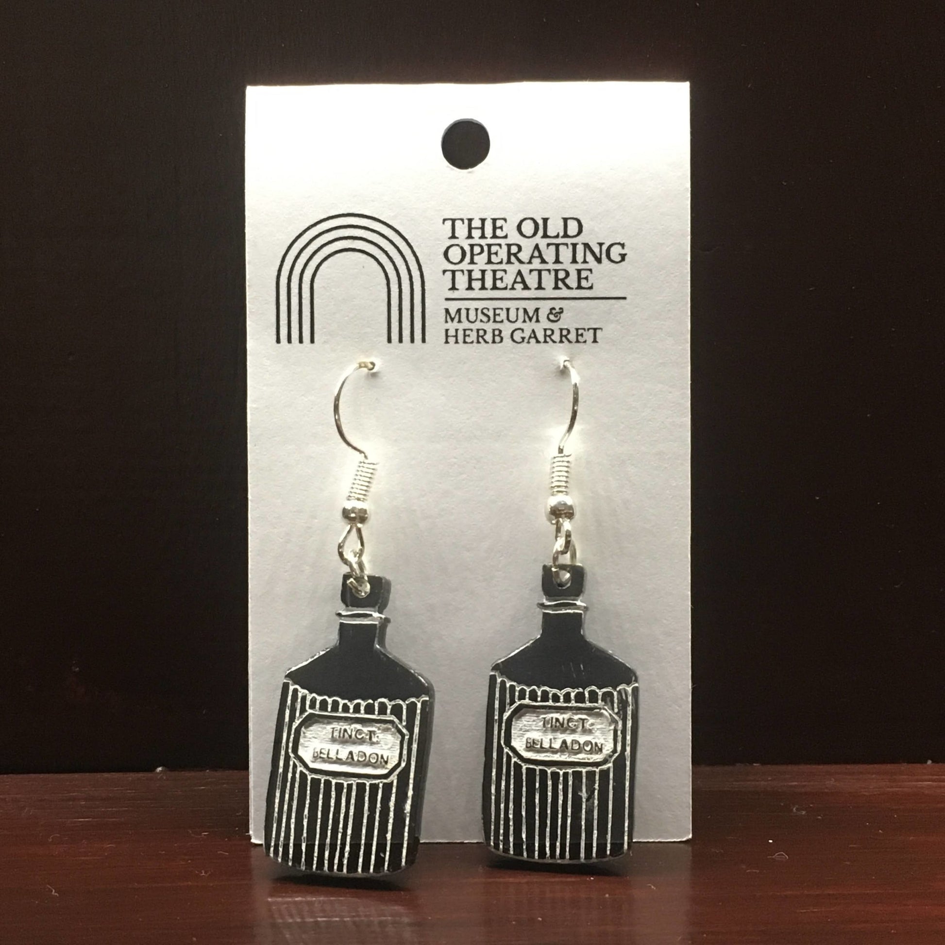 Pair of black dangle earrings in the shape of a bottle with the label 'tinct. belladon'