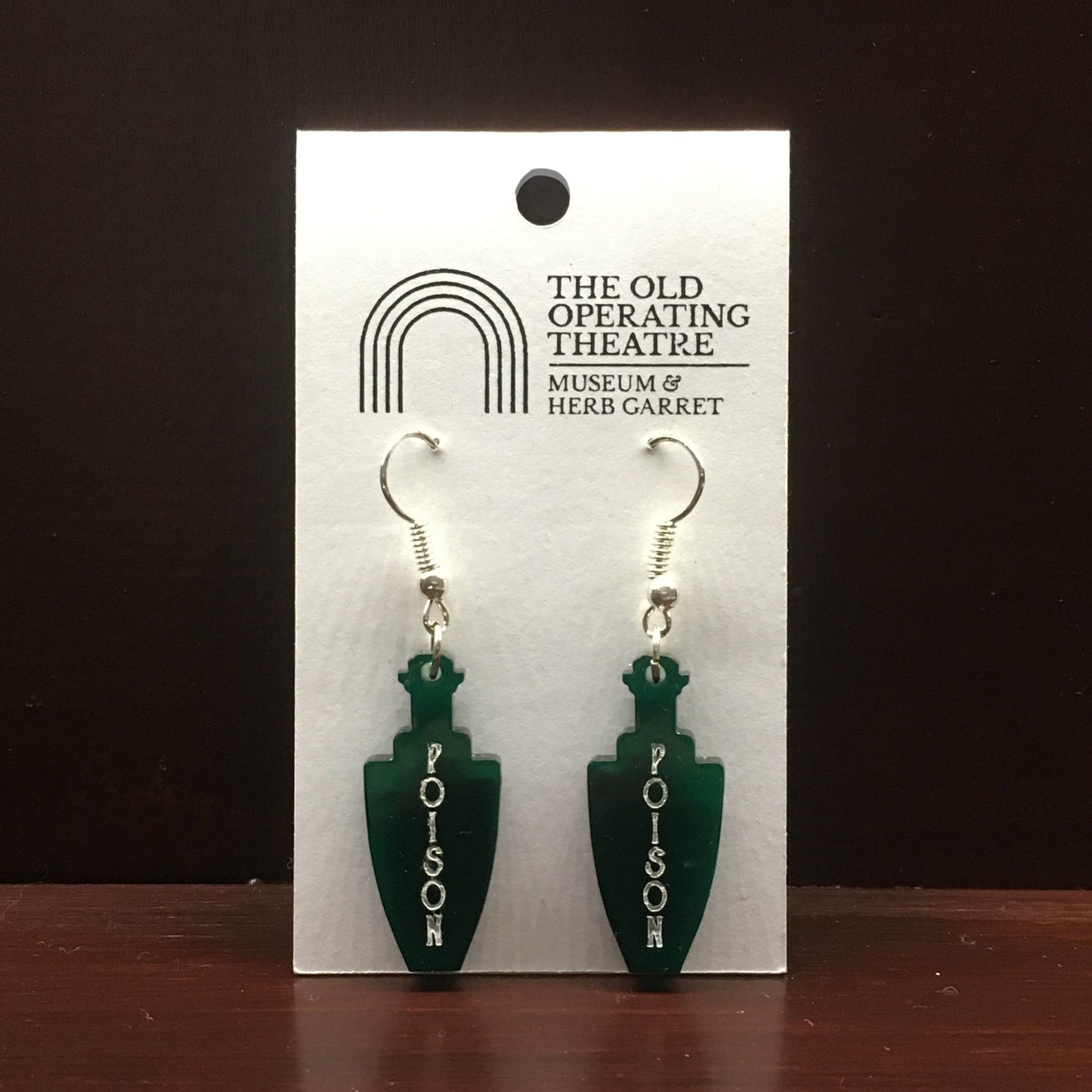 Pair of green dangle earrings in the shape of a bottle. 'Poison' is written vertically on the bottle.