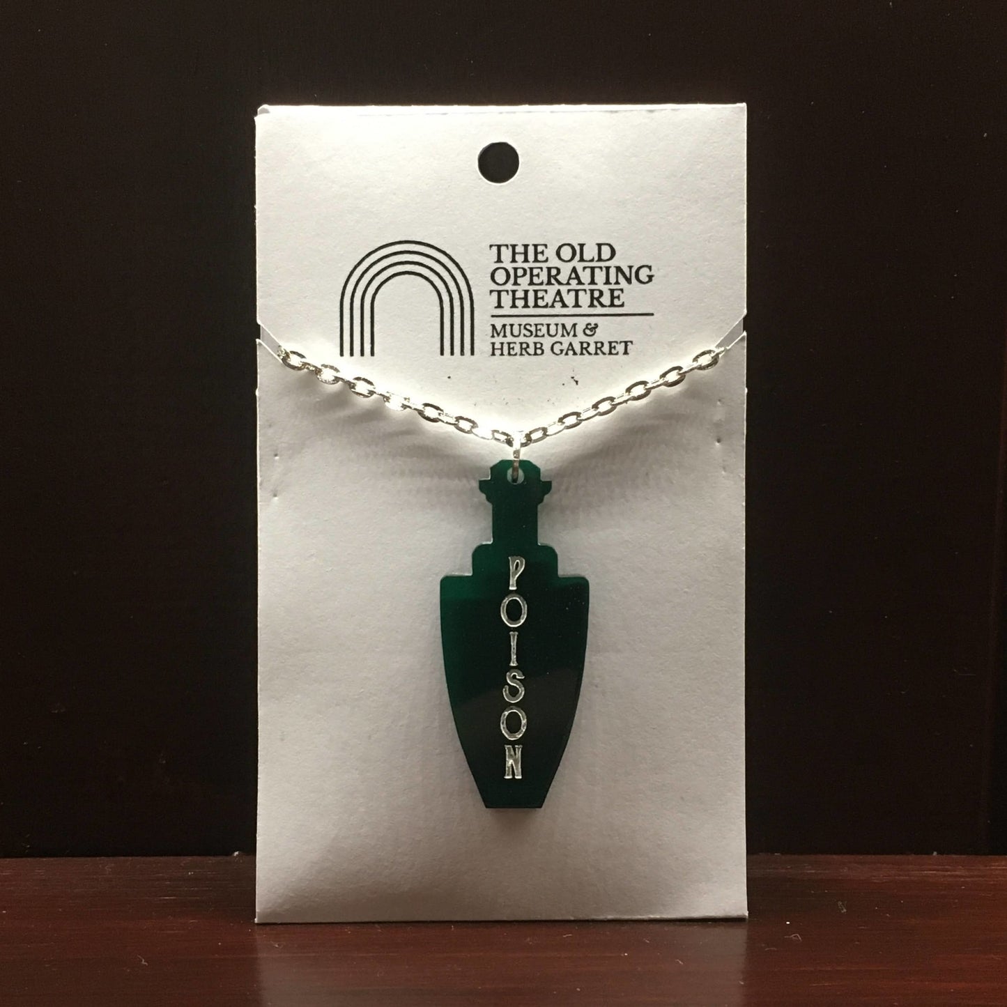 Green pendant in the shape of a bottle, with 'poison' written vertically on the front. On a metal chain.
