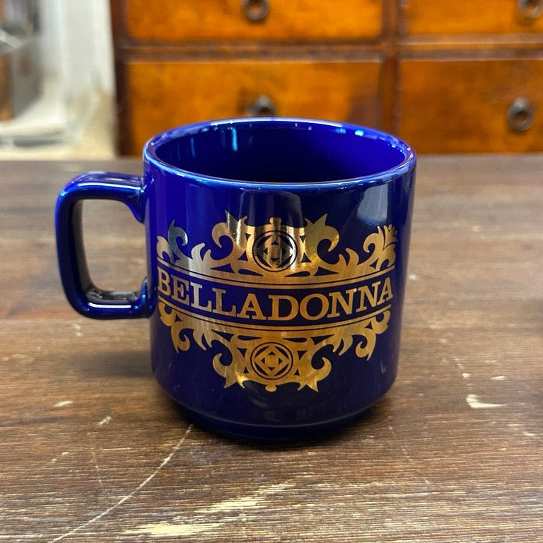 Blue mug with gold 'Belladonna' logo on a wooden surface