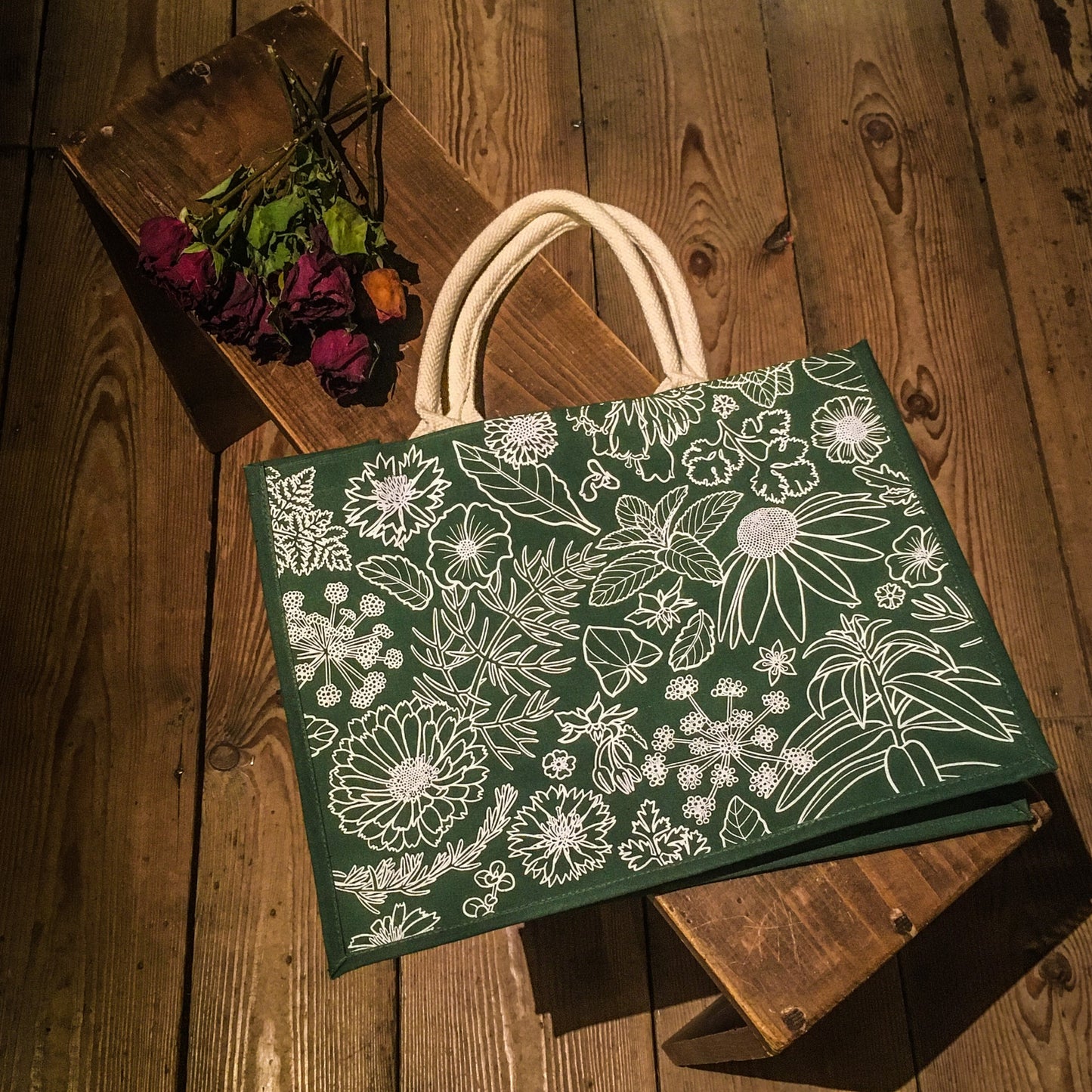 Green bag with white outline decoration of flowers and leaves, placed on a wooden bench next to some dried roses