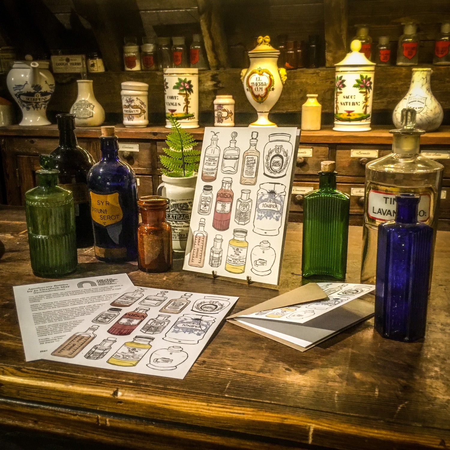 Two prints and a greetings card featuring the apothecary bottles design on the museum's wooden apothecary counter, next to some of the glass apothecary bottles in the museum's collection