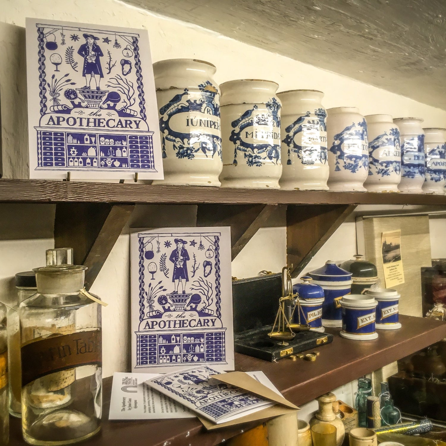 Two prints and a greetings card featuring the blue and white apothecary design, placed on shelves in the museum next to a row of blue and white apothecary jars