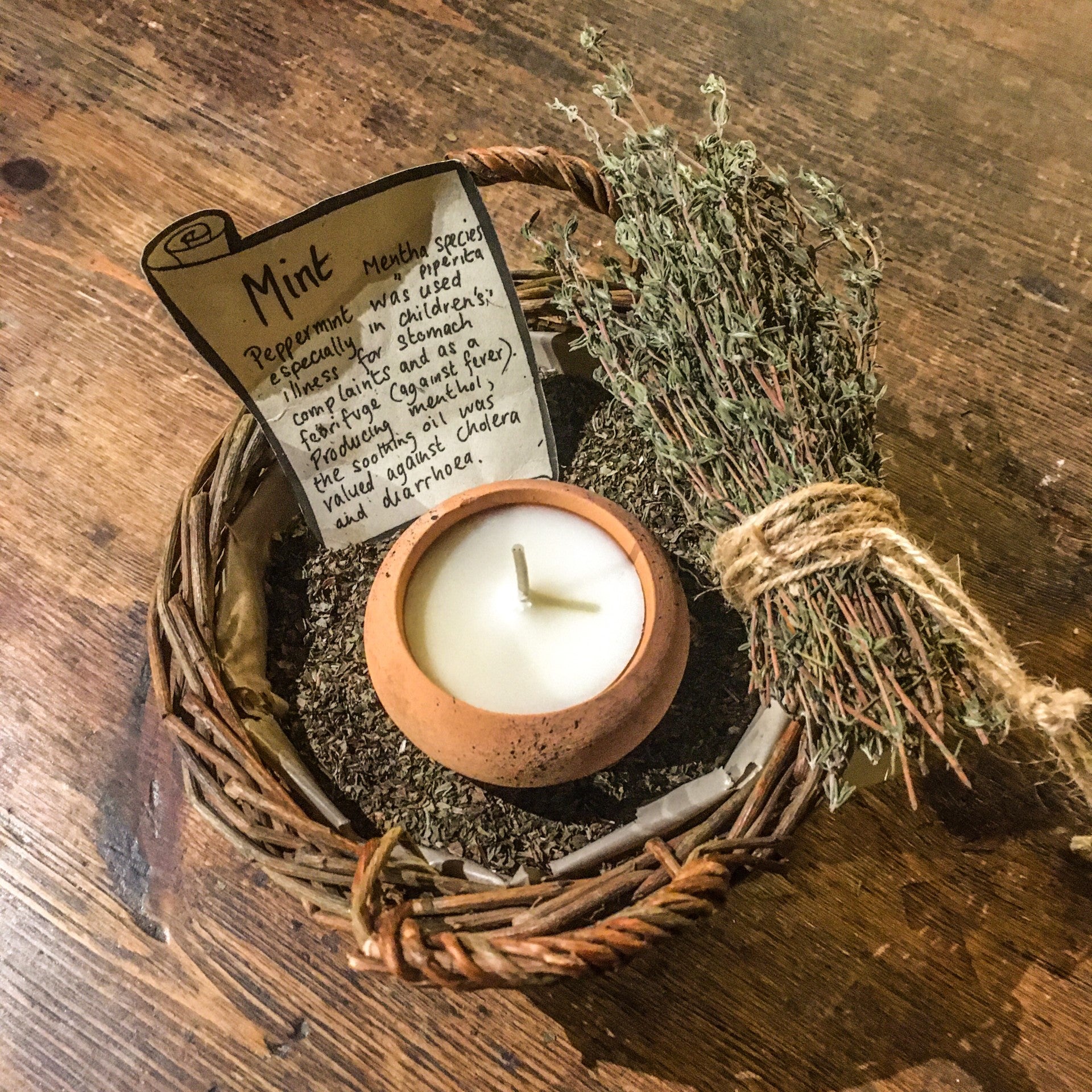 A terracotta tealight placed in a basket of dried mint next to a bundle of thyme sprigs tied with brown string