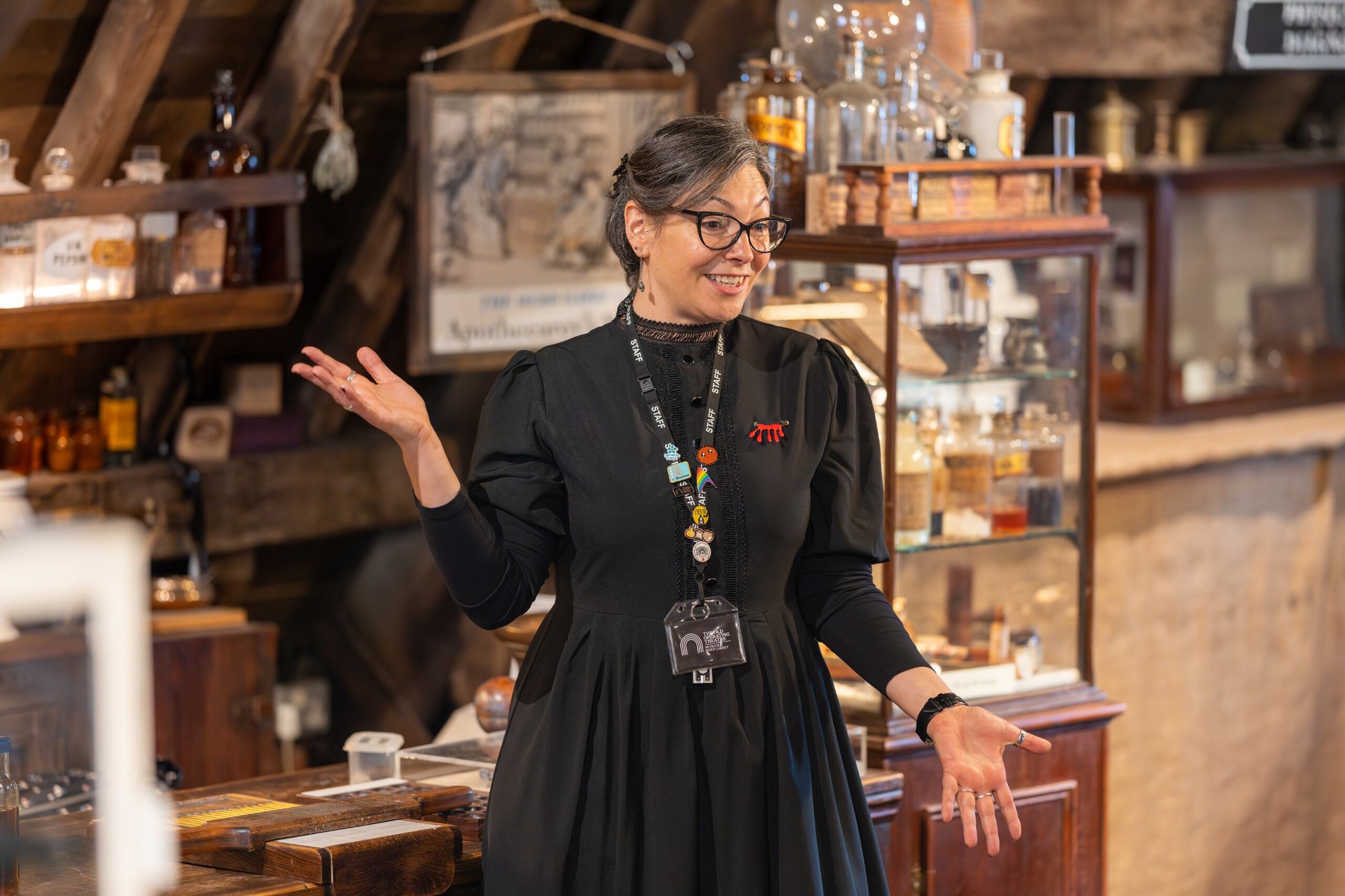 A woman in a black dress and glasses stands in the museum, gesturing with her hands