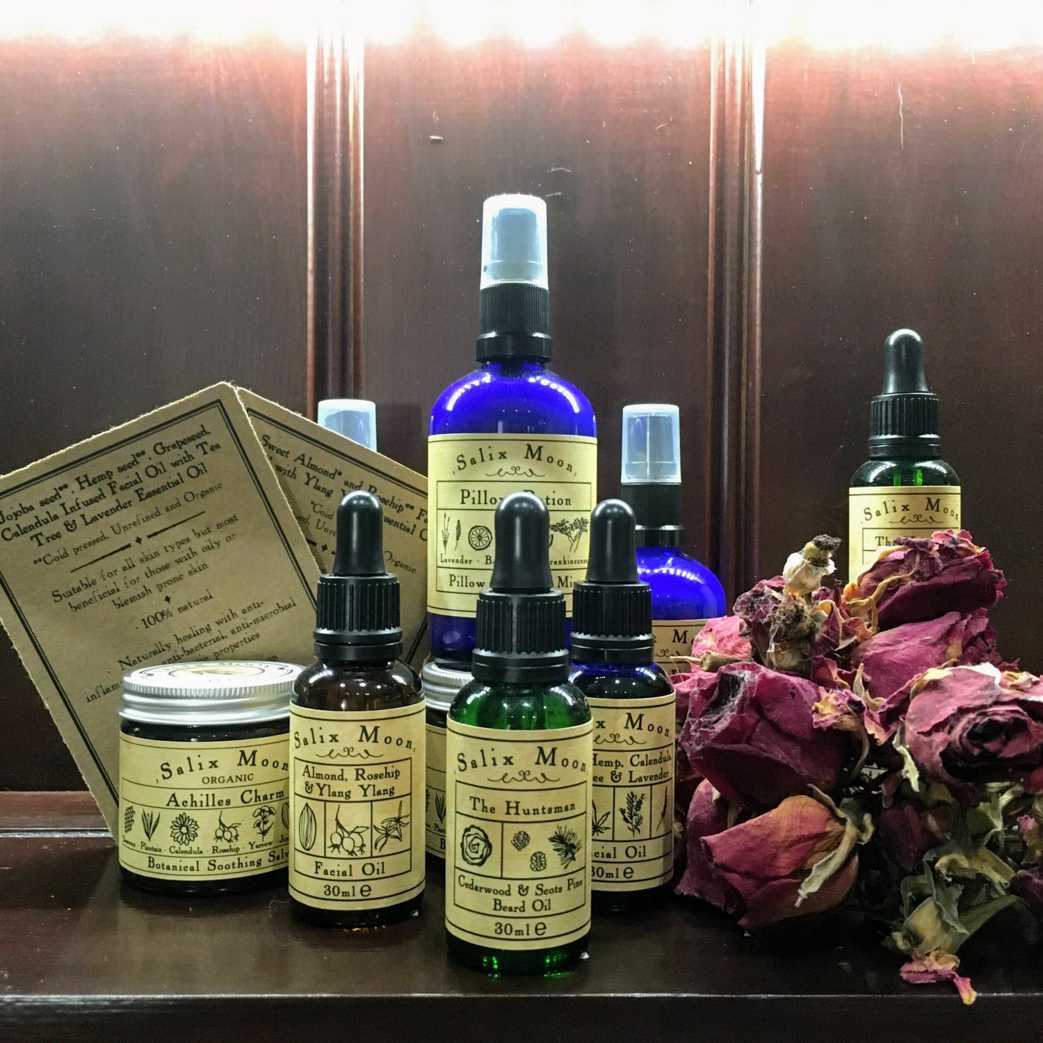Bottles of pillow potion, beard oil and facial oil, and a jar of Achilles Charm salve, next to dried roses
