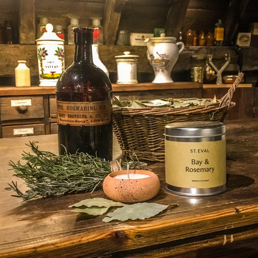 A terracotta candle and a bay and rosemary candle in a tin, on the museum's apothecary counter next to some bay leaves, a bundle of rosemary sprigs and an old brown glass bottle with 'rosmarini' visible on the worn label