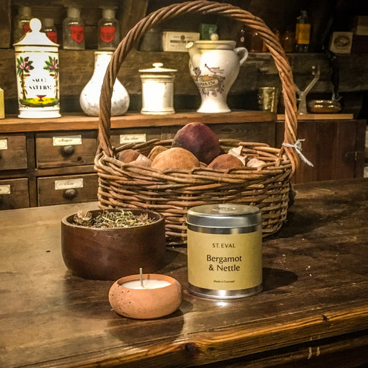 A terracotta candle and a bergamot and nettle candle tin on the museum's apothecary counter, next to a wooden bowl of herbs and a wicker basket of dried fruit
