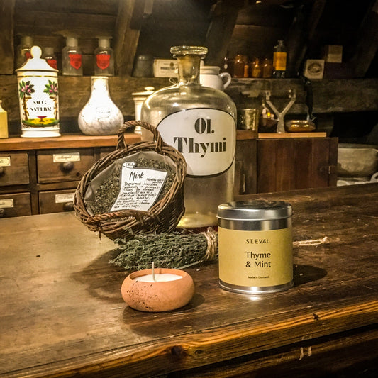 A terracotta tealight and a thyme and mint candle tin on the museum's apothecary counter, next to a bunch of dried thyme, a basket of dried mint and a large glass bottle labelled 'Ol. Thymi'