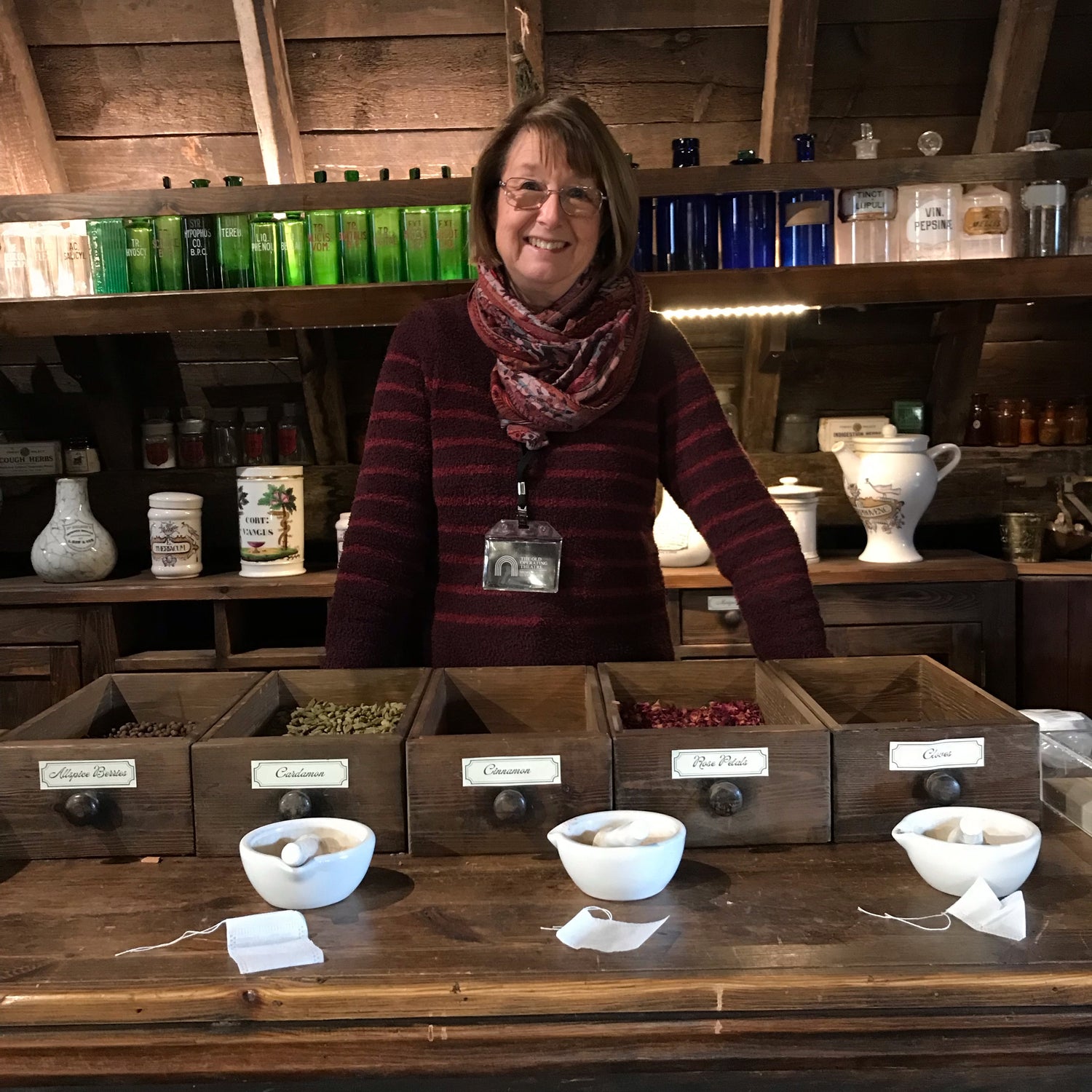 A smiling woman standing at the apothecary counter with drawers of dried herbs and small pestles and mortars in front of ehr