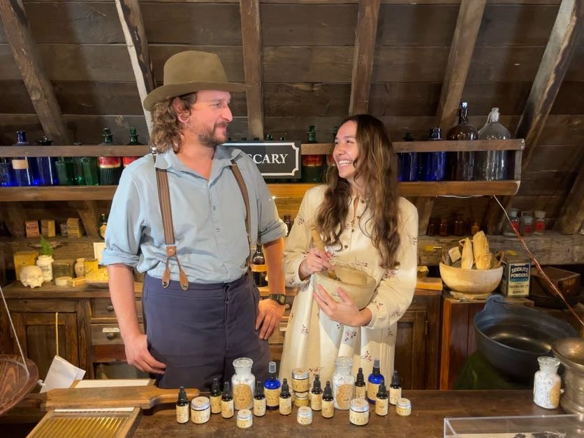 A woman using a pestle and mortar smiles at a man as they stand behind the museum's apothecary counter, on which are a range of Salix Moon products