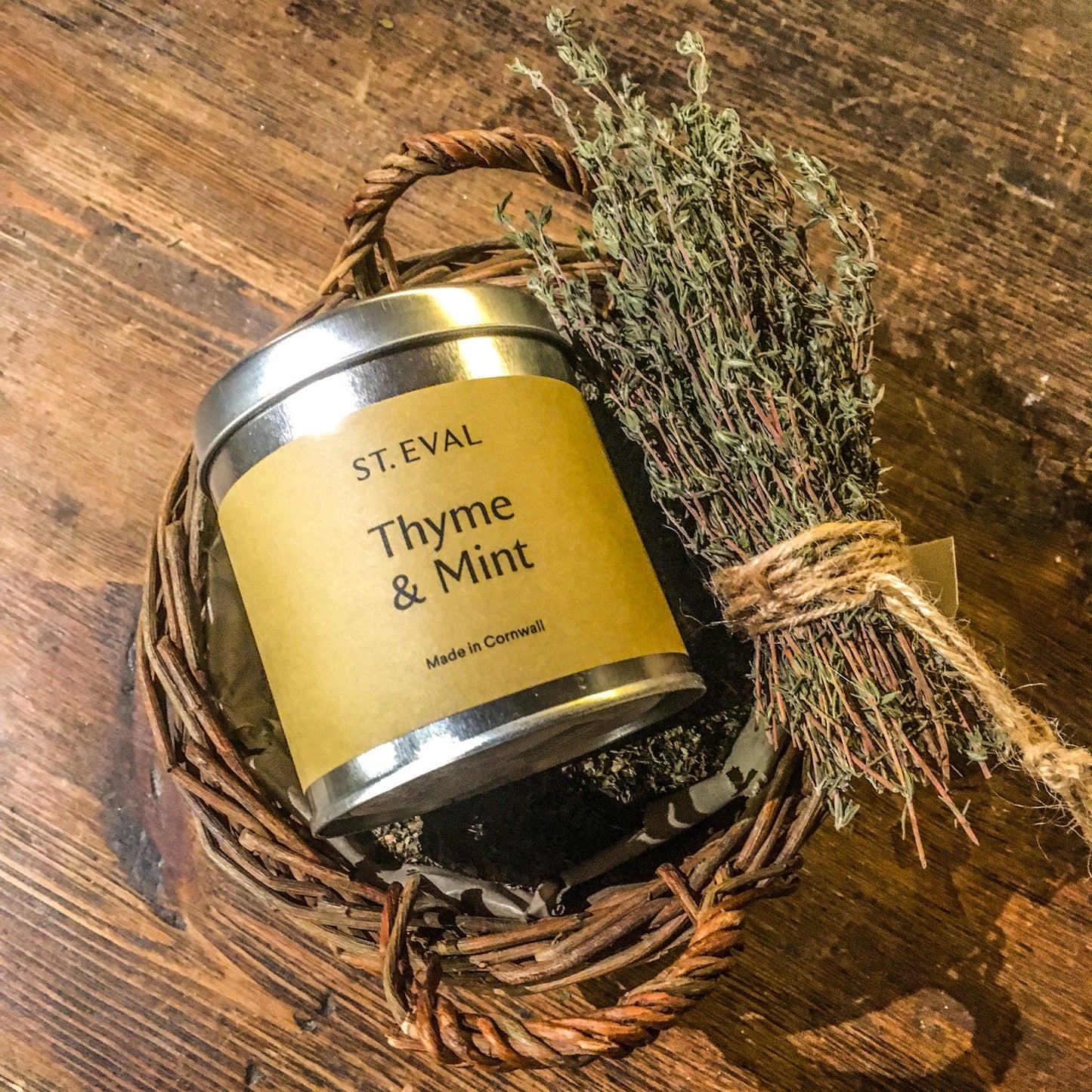 A thyme and mint candle tin placed in a basket of dried mint next to a bundle of thyme sprigs tied with brown string