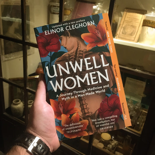 The Unwell Women book being held up in front of a display in the museum featuring objects from women's medicine and domestic health