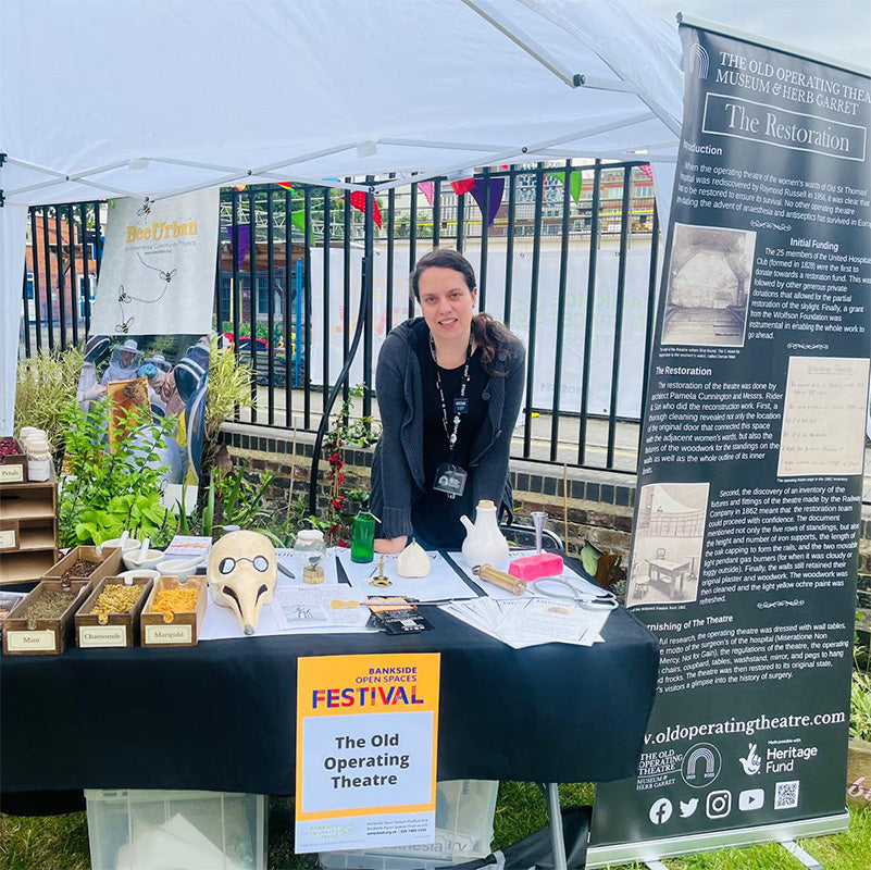 Colour photo of a woman wearing a museum lanyard standing behind a table under a large gazebo; on the table are boxes of herbs, sheets of paper and objects from the museum, including a plague doctor mask; next to the table is a pull-up banner about the museum