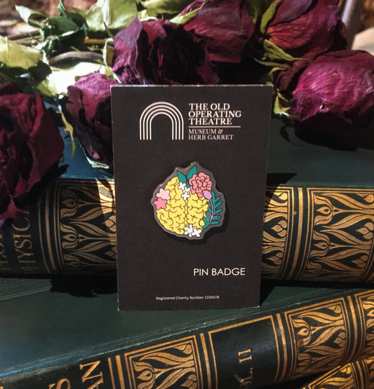 A pin with a yellow brain decorated with pink, white and green leaves and flowers, placed on a set of hardback books on top of which are dried roses