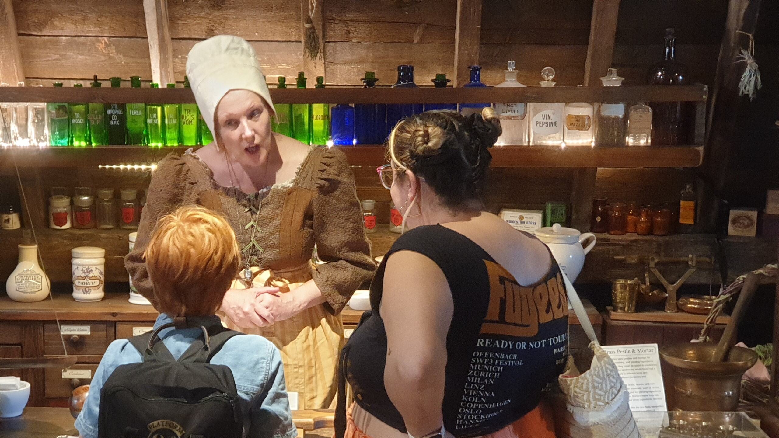 A woman in historical costume stands behind the apothecary counter and speaks to an adult and child visitors