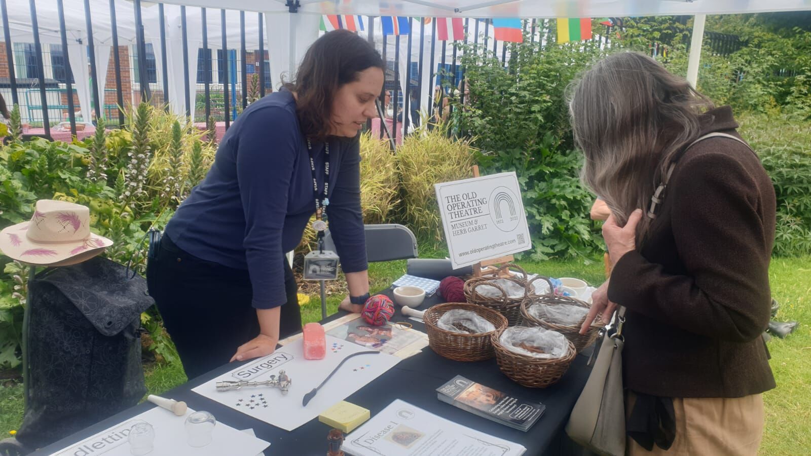 A museum representative standing behind a table outdoors, talking to a visitor about items in baskets