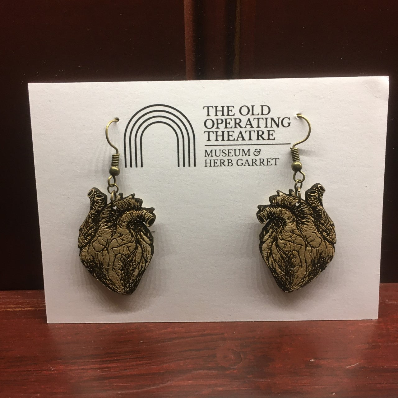 A pair of wooden earrings in the shape of an anatomical heart, dangling from a white card with the museum logo