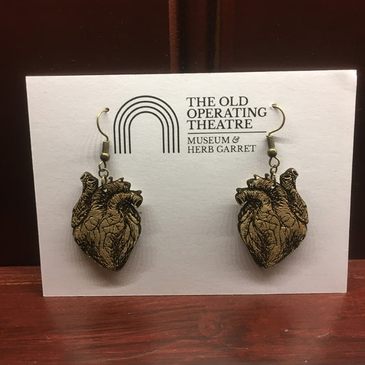 A pair of wooden earrings in the shape of an anatomical heart, dangling from a white card with the museum logo