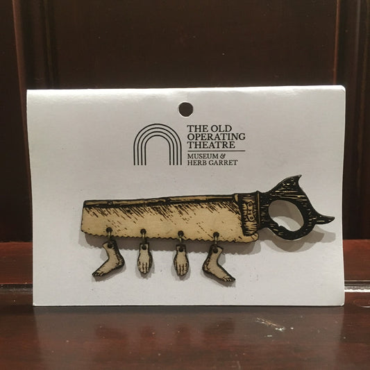 A wooden brooch in the shape of a saw, with two hands and two feet dangling from it