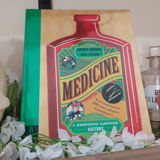 The book Medicine A Magnificently Illustrated History propped up next to a glass medicine bottle and with some white flowers lying across the bottom