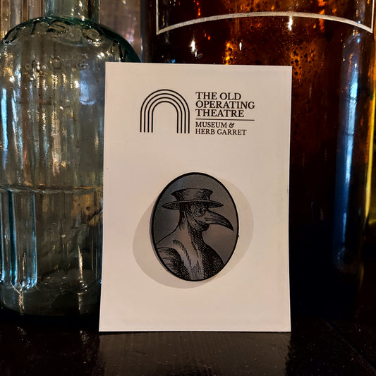 An oval brooch with a plague doctor design in black and silver, on white card with the museum's logo, propped against some of the museum's glass bottles