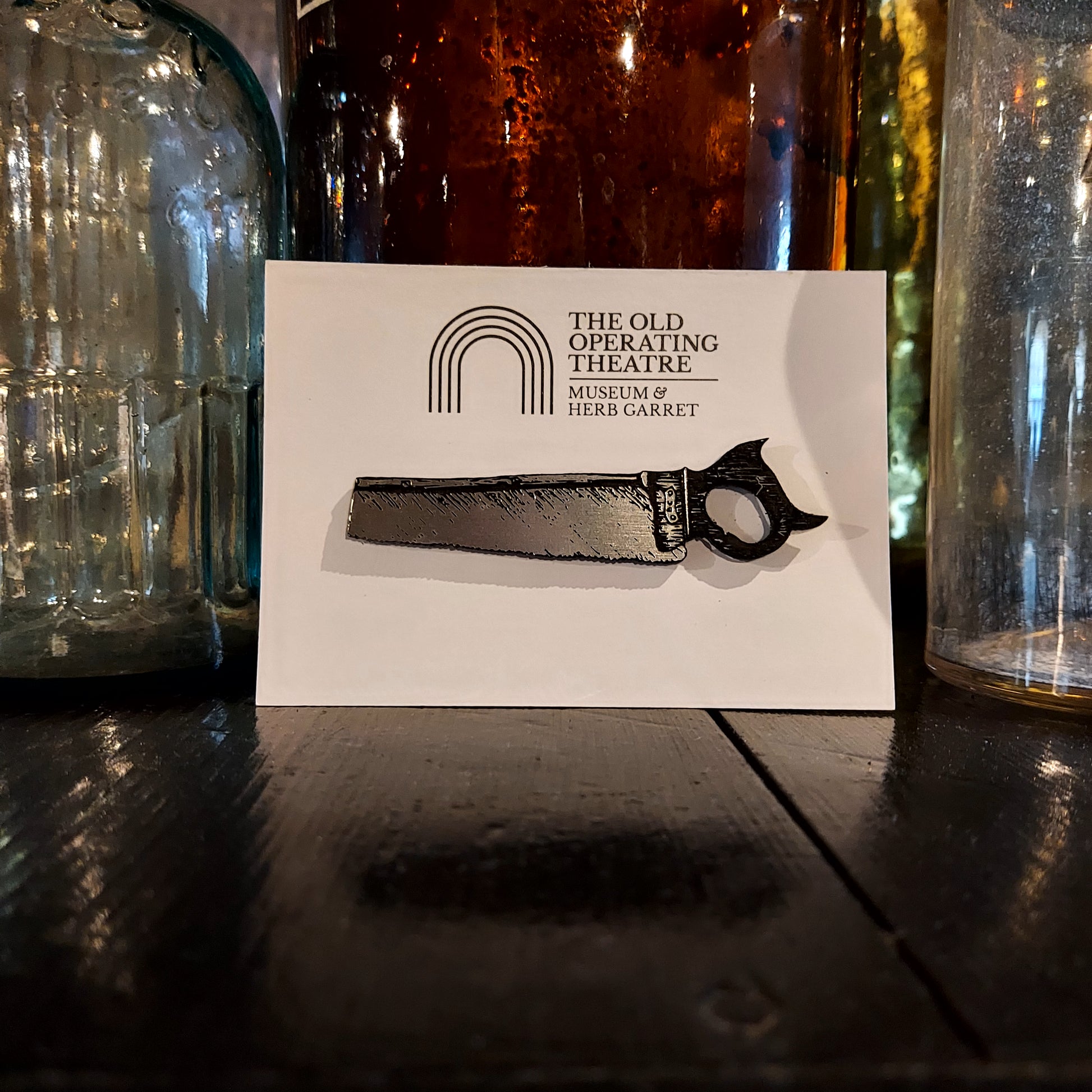 A silver-coloured brooch in the shape of a saw on a piece of white card with the museum's logo, placed against a selection of glass bottles in the museum