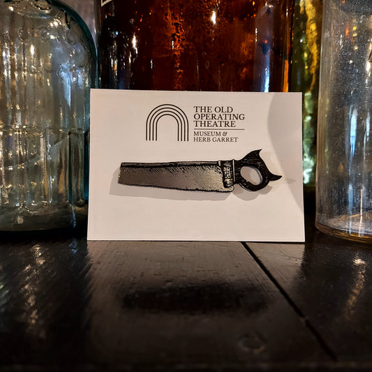 A silver-coloured brooch in the shape of a saw on a piece of white card with the museum's logo, placed against a selection of glass bottles in the museum