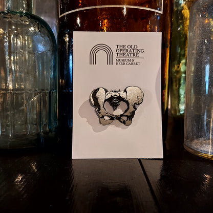 A silver-coloured brooch in the shape of the pelvic bones on a piece of white card with the museum's logo, propped against a selection of the museum's glass bottles