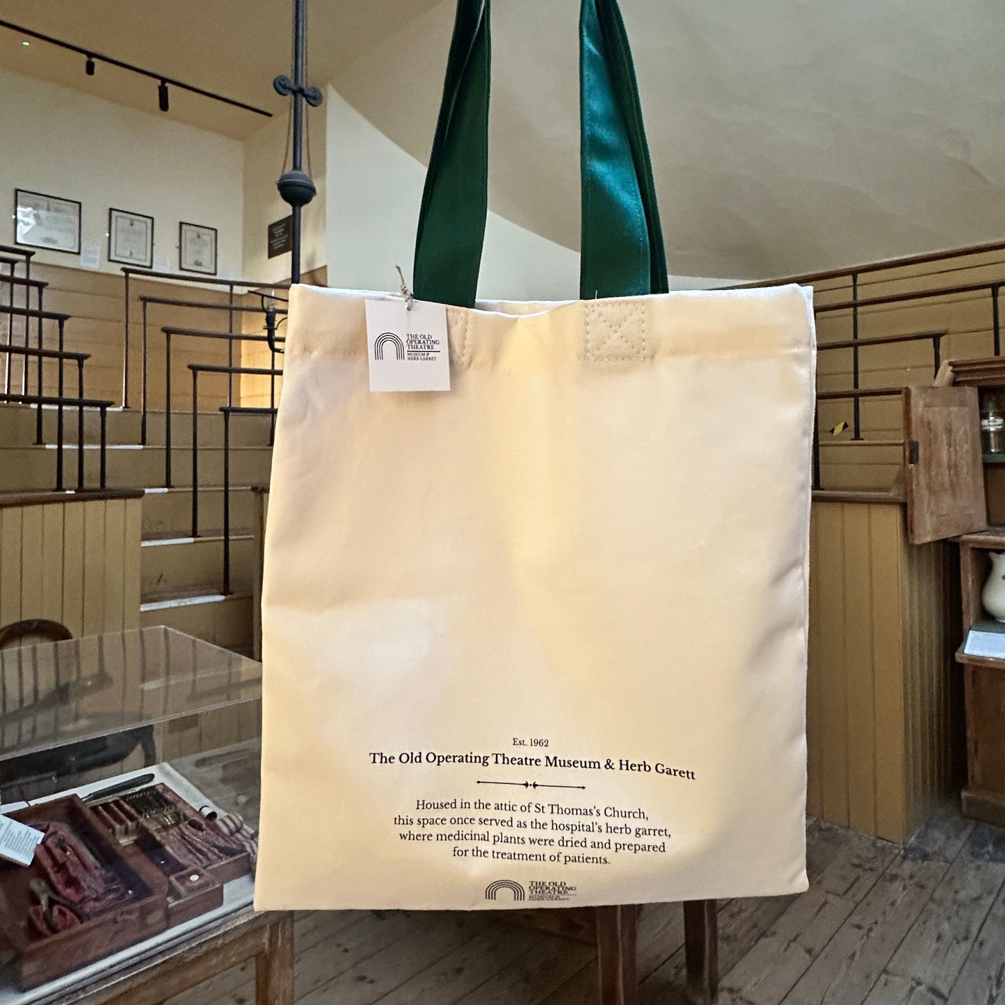 A cream canvas tote bag with dark green shoulder straps, held up by a hand inside a modern gallery or museum space. The bag features printed black text and a small white logo near the top left.
