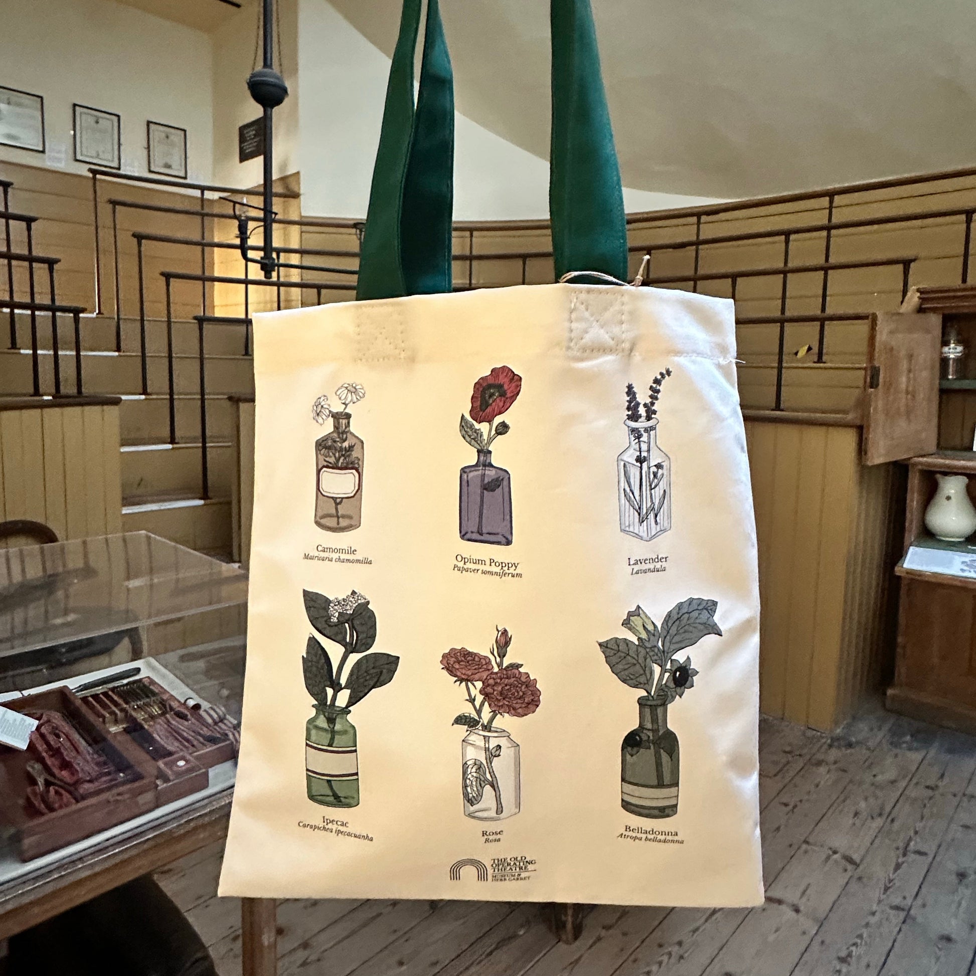 A cream canvas tote bag with dark green handles, featuring illustrations of six different perfume bottles and labels, held up by a hand inside the old operating theatre 