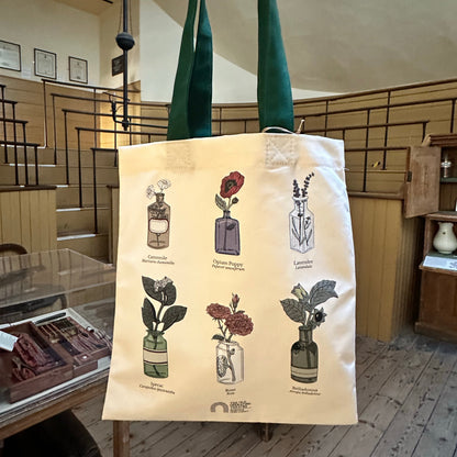 A cream canvas tote bag with dark green handles, featuring illustrations of six different perfume bottles and labels, held up by a hand inside the old operating theatre 