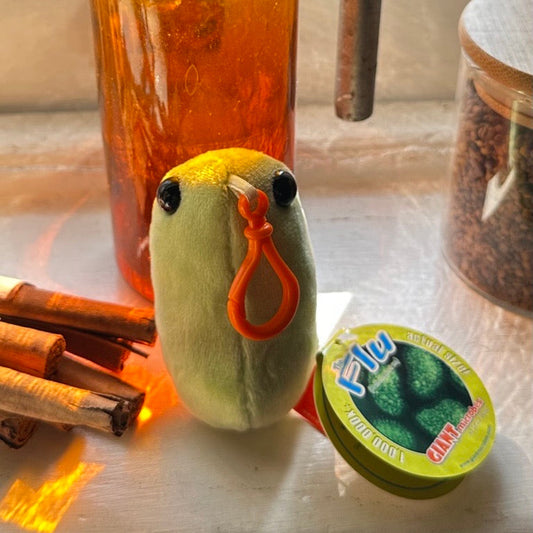 Small green plush toy with an orange clip on a surface with various items including a bottle and cinnamon sticks.