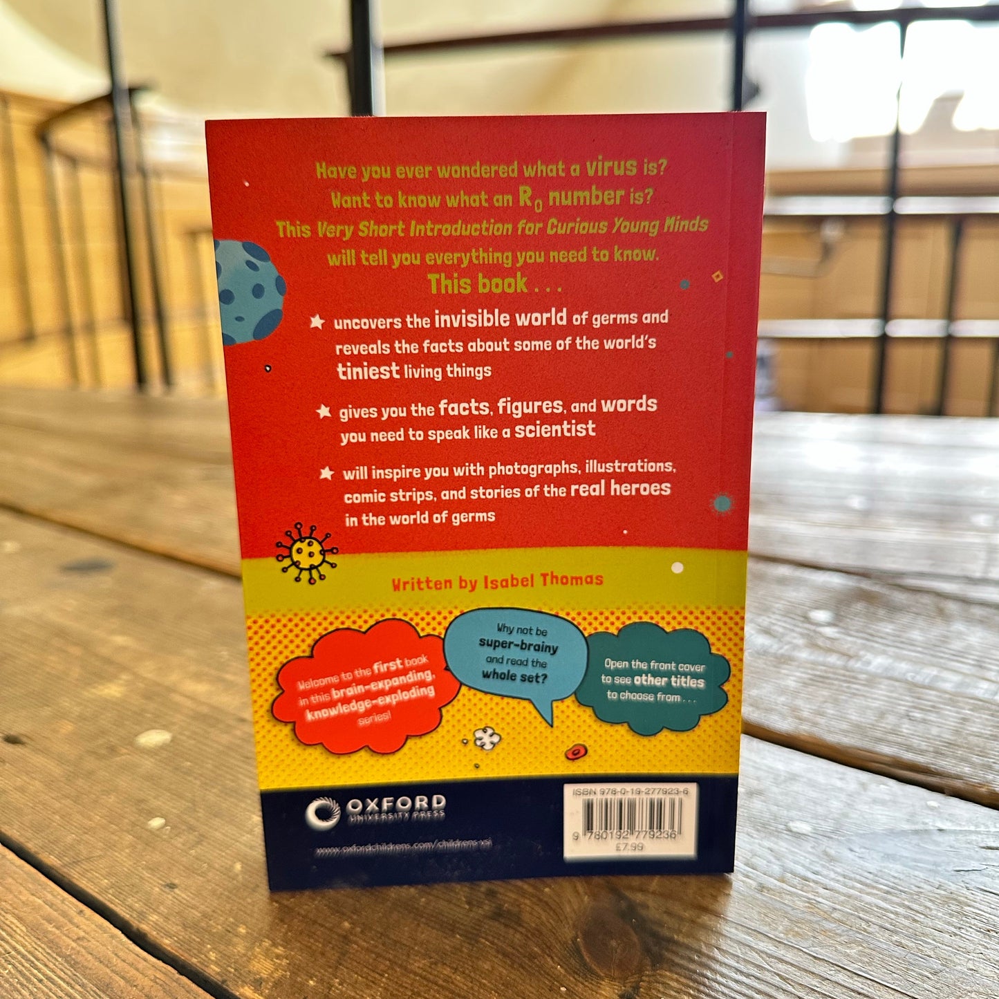 The back cover of the Oxford This Week Short Infraction for Durable Young Minds book, featuring a colorful design with illustrations and text, photographed on a wooden table.