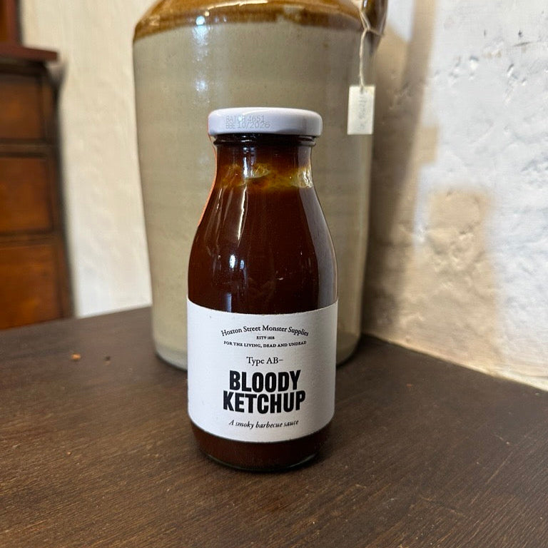 Bottle of Bloody Ketchup on a wooden surface with a large ceramic jar in the background.
