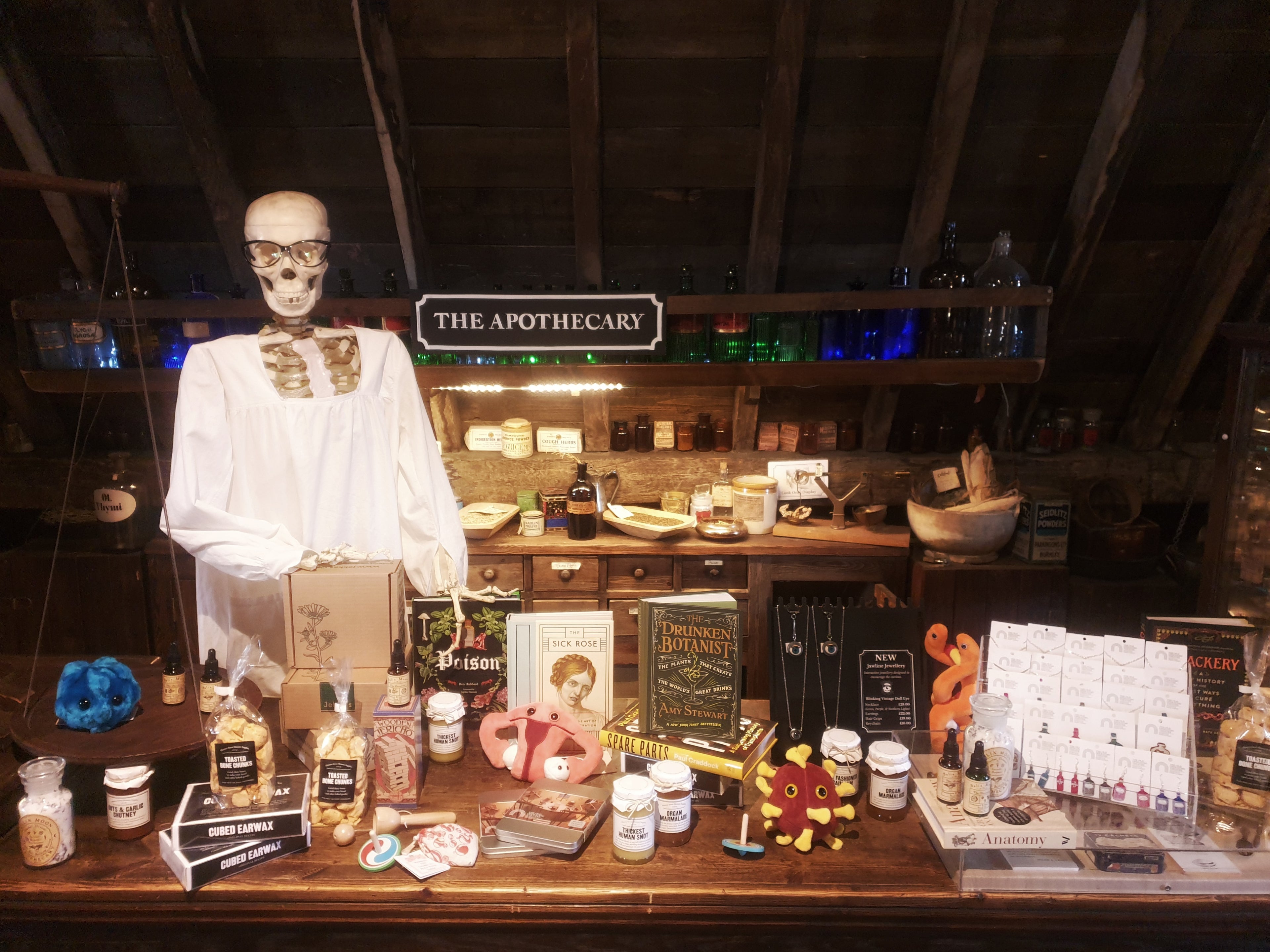 A skeleton wearing glasses and a white gown behind the apothecary counter, on which is a splendid array of shop products, including books, jewellery, plush toys, jam and toiletries